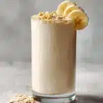 Un grand verre de smoothie protéiné banane avoine onctueux, garni d'une tranche de banane et de quelques flocons d'avoine, sur un fond clair.