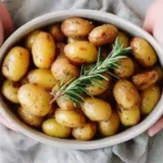 Gros plan sur un bol de pommes de terre grenaille au four dorées, garnies de romarin frais et tenues par des mains.