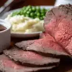 Rôti de bœuf au four mi-saignant, tranché, avec sauce, purée et petits pois sur une planche en bois.